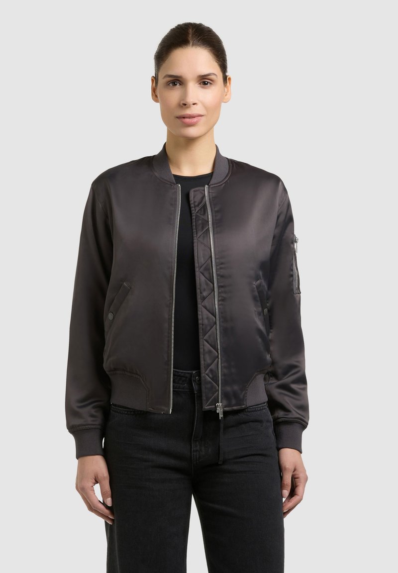 Femme debout, portant une veste bomber noire sur une chemise noire et un pantalon noir, devant un fond clair uni.