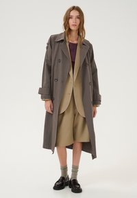 Dlouhý šedý trench coat s dvojitým zapínáním, velkými černými knoflíky a rukávy se svršky, nošený přes béžový oblek a hnědé mokasíny.
