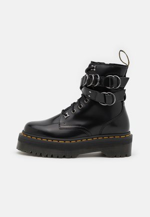 Black shiny platform boot with thick rubber sole, yellow stitching, lace-up front, and double buckle straps around the ankle.