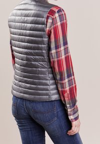 Šedý péřový vest s horizontálním prošíváním v kombinaci s košilí v kostičkovaném vzoru v červené, modré a zelené barvě. Džíny z rifloviny doplňují celkový vzhled.