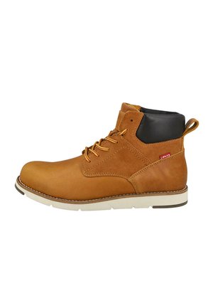 Botín de cuero y ante color canela con cuello acolchado negro, cordones delanteros, suela blanca y pequeña etiqueta roja en el lateral.