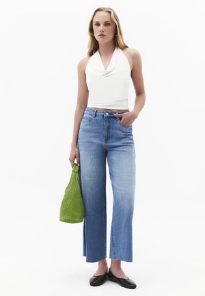 Junge Frau trägt weißes Neckholder-Top, hochtaillierte Jeans mit weitem Bein, schwarze flache Schuhe, hält grüne geflochtene Handtasche, steht mit einer Hand in der Tasche.