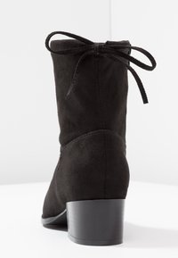 Bottine en daim noir avec une finition texturée, bout arrondi et petit talon bloc. Présente un détail de nœud à l'arrière pour un design supplémentaire.