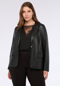 Vrouw in een zwarte leren blazer over een zwarte kanten top en zwarte broek, die zelfverzekerd naar de camera kijkt tegen een effen achtergrond.