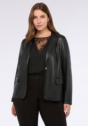 Vrouw in een zwarte leren blazer over een zwarte kanten top en zwarte broek, die zelfverzekerd naar de camera kijkt tegen een effen achtergrond.