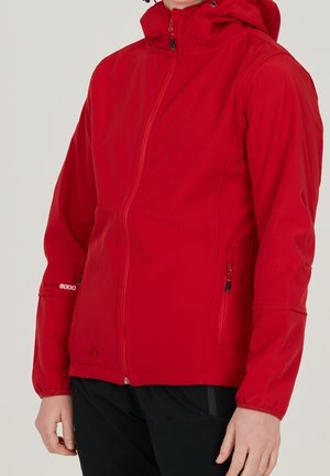 Person trägt eine rote Jacke mit Kapuze und Reißverschlusstaschen vorne und an den Seiten, kombiniert mit einer schwarzen Hose, gezeigt von der Brustmitte bis zur Hüfte.