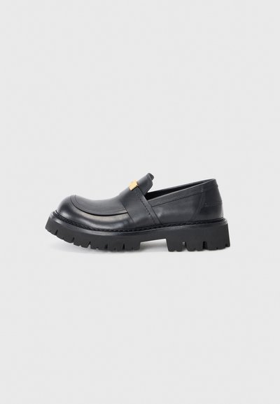 Schwarzer Slip-On-Schuh aus Leder mit abgerundetem Zehenbereich, genoppter Sohle und goldfarbenem Akzentriemen. Verfügt über eine klobige, robuste Außensohle für gute Traktion.