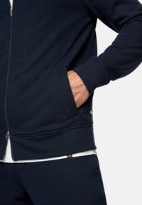 Felpa con zip di colore blu navy, caratterizzata da una texture morbida, tasche laterali, polsini a coste e un accento logo discreto vicino all'orlo. Indossata sopra una maglietta bianca.