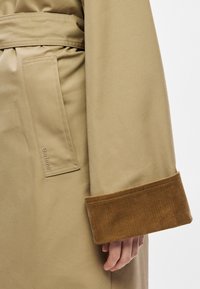 Beežika värvi trentšmantel sametpihlaga, küljetasku ja reljeefse brändilogoga. Sile kangapind, vööga disain.