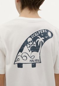 Bílý bavlněný tričko s tmavě modrou grafikou surfařské desky se skull, palmami, horami a nápisy "SCALPERS" a "PURÁ VIDA."