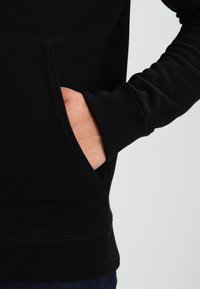 Felpa nera con tasca a marsupio, polsini e orlo a costine. Il materiale sembra morbido con una texture liscia e dettagli di cucitura minimi.