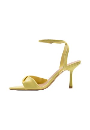 Sandalo giallo con tacco alto, sottile cinturino alla caviglia e fascia frontale attorcigliata, punta aperta e tacco a spillo su sfondo bianco.