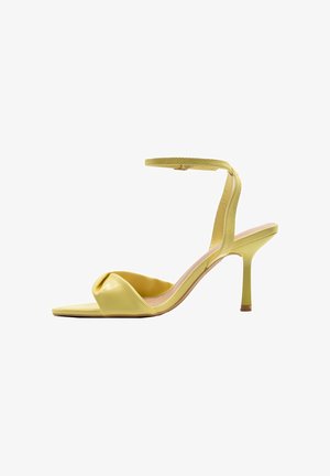 Sandalo giallo con tacco alto, sottile cinturino alla caviglia e fascia frontale attorcigliata, punta aperta e tacco a spillo su sfondo bianco.
