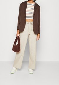 Person klädd i en brun blazer, randig crop top, beige vida byxor, vita sneakers, som håller en brun fluffig handväska mot en neutralt bakgrund.