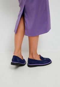 Laivastonsinisen mokka-liukupohjaiset kengät, joissa on violetti koristereuna ja musta pohja. Mallilla on päällään violetti hame, jossa on sivuavoinne, joka paljastaa osan jalasta.
