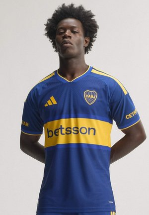 Mann mit Afro-Frisur trägt ein blau-gelbes Boca Juniors Fußballtrikot mit Sponsorlogos und steht vor einem schlichten Hintergrund.