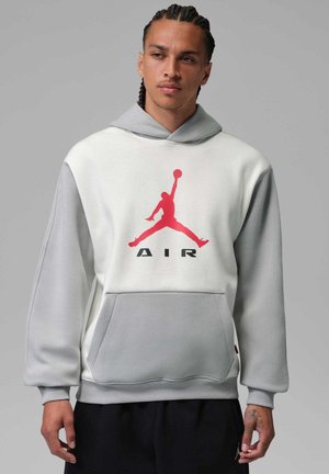 Mand med flettet hår iført en grå og hvid hættetrøje med en rød basketballspillerlogo og sorte bukser mod en ensfarvet grå baggrund.