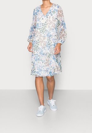 Vrouw draagt een knielange, lange mouwen bloemenjurk met blauwe en pastelpatronen, gecombineerd met lichtblauwe sneakers.