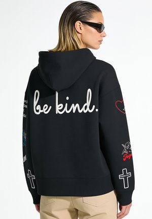 Femme aux cheveux lissés en arrière portant un sweat à capuche noir avec le texte "be kind." dans le dos et des motifs graphiques sur les manches, pantalon beige et lunettes de soleil.