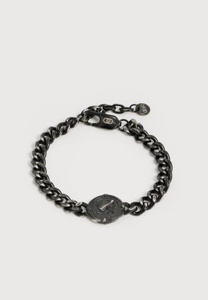 Čierny kovový reťazový náramok s okrúhlym gravírovaným príveskom v tvare mince a karabínkou s malou značkovou visačkou.