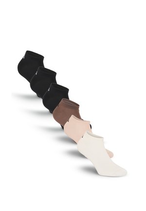 Personne noire aux cheveux courts se tient sur des marches en béton, portant un t-shirt blanc, un short blanc et des chaussettes blanches devant un mur en béton.