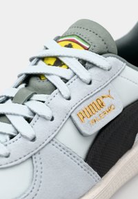 Heledad hallid tossud, millel on nubukist ja nahast materjalid. Hallid accendid, valged paelad ja kullast logo silt "PUMA PALERMO."