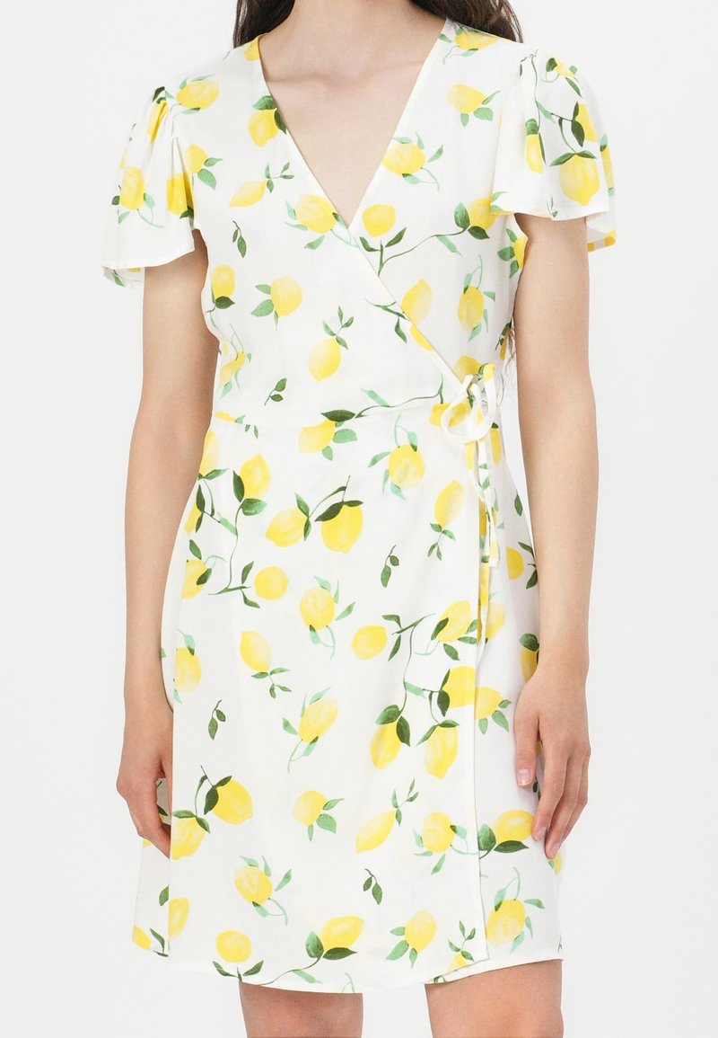 Robe portefeuille blanche imprimée de citrons jaunes et de feuilles vertes, manches courtes à volants et détail noué sur le côté, portée par une personne debout.