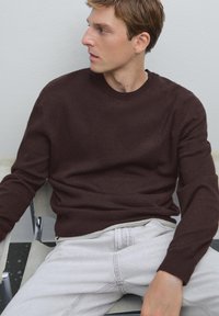 Maglione marrone a maglia con scollatura rotonda e maniche lunghe, abbinato a jeans grigi chiari, seduto su una sedia di metallo contro un muro neutro.