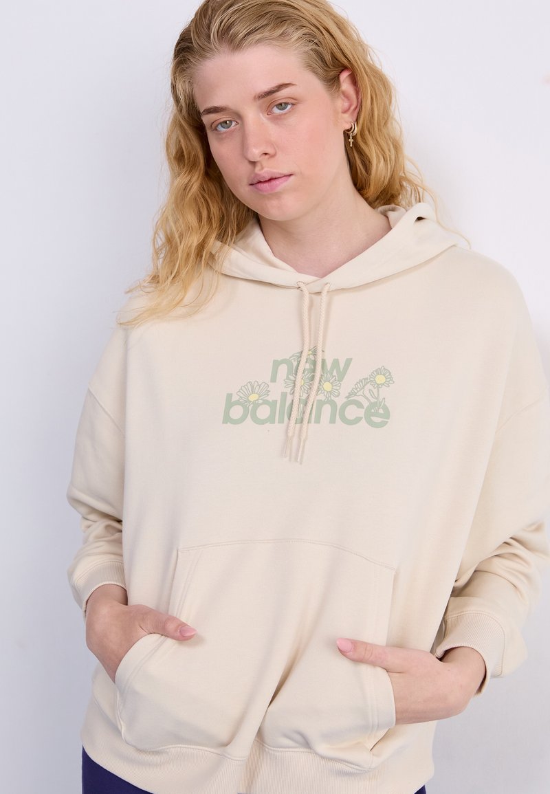Beige Hoodie mit einer Fronttasche und Kapuze mit Kordelzug. Mit "New Balance" Grafik und floralen Akzenten in grün. Weicher Stoff.