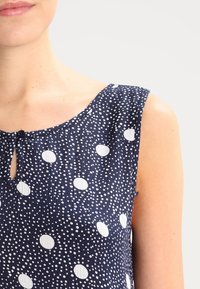Marineblauwe mouwloze top met witte stippen, met een keyhole-opening en een textuurstof, die het lichte ontwerp benadrukt.