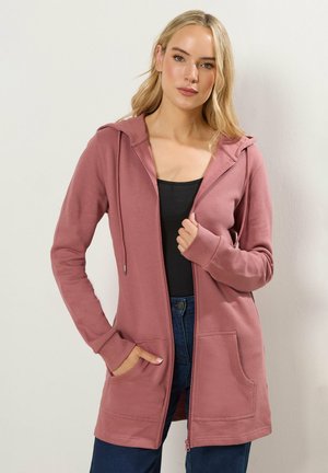 Frau trägt einen langen, staubig rosa Reißverschluss-Hoodie mit Taschen, schwarzen Oberteil und blauen Jeans, steht vor einer schlichten hellen Wand.