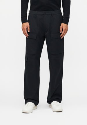 Άνδρας που φοράει μαύρο παντελόνι cargo με τσέπες με καπάκι και λευκά αθλητικά παπούτσια, στέκεται μπροστά σε απλό φόντο.