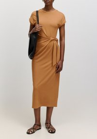 Robe midi jaune moutarde, manches courtes, détail noué sur le côté. Associée à un sac à épaule noir et des sandales noires à lanières. Tissu lisse.