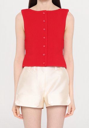 Haut rouge sans manches boutonné associé à un short crème taille haute avec poches à rabat à l’avant, porté par un mannequin debout.