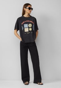 Schwarzes Grafik-T-Shirt mit kurzen Ärmeln und lockerer Passform, das einen bunten "Nimm eine Sonnenpause"-Druck zeigt, kombiniert mit schwarzen weiten Hosen.