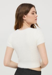 Maglione bianco a maniche corte realizzato in un materiale a maglia pelosa con un design leggermente accorciato. La texture appare morbida e accogliente, con dettagli minimi.