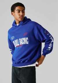 Νεαρός άνδρας που φοράει μπλε φούτερ με κουκούλα με λευκά και κόκκινα διακριτικά αγωνιστικού τύπου, μαύρο τζιν, κοιτάζοντας προς τα πλάγια με φόντο απλό γκρι ανοιχτό.