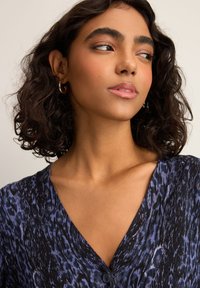 Haut à col en V en tissu noir texturé avec un imprimé léopard bleu. Dispose d'une fermeture à bouton et d'un décolleté doux et arrondi. Cheveux coiffés en boucles lâches.