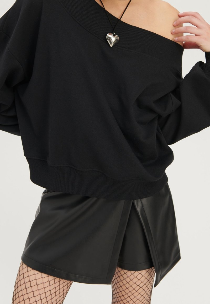 Femme portant un pull noir épaules dénudées, un short jupe portefeuille en simili cuir noir, des collants résille et un collier pendentif en argent en forme de cœur.