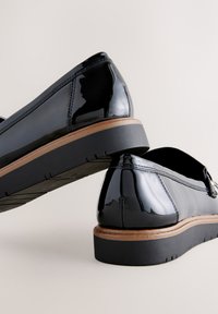 Mocassins en cuir verni noir avec une semelle empilée marron et des détails de couture subtils, présentant un embout arrondi et une texture lisse.