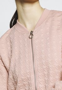 Donna che indossa una giacca bomber texturizzata rosa chiaro con una cerniera argentata e un anello di estrazione, capelli raccolti, su uno sfondo semplice.