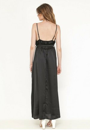 Robe maxi noire en satin sans manches avec une taille froncée. Présente une texture lisse et une silhouette longue et fluide. Des chaussures à talons hauts blanches sont visibles.