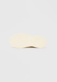 Podplat čevlja iz gume kremne barve z teksturiranim vzorcem in blagovno znamko, s gladko površino in rahlo dvignjenim profilom.