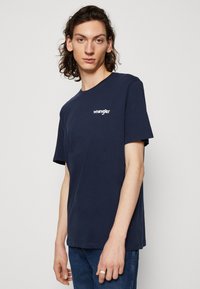 Temno modra bombažna majica s kratkimi rokavi, na levi strani prsnega koša z belim napisom "Wrangler". Preprost dizajn z okroglim izrezom.