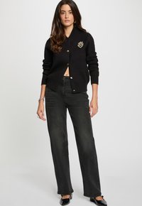 Cárdigan de punto negro con cuello redondo y botones dorados, que presenta un parche bordado en el pecho; combinado con jeans oscuros de cintura alta.