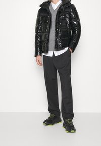 Zwarte glanzende pufferjack, grijze trui, witte shirt, grijze broek en zwarte sneakers met neongroene accenten. Modern, gelaagd outfitontwerp.