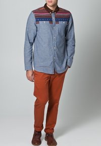 Camisa abotonada de tela azul claro con canesú estampado en rojo, azul y marrón; combinada con pantalones naranjas y zapatos marrones.