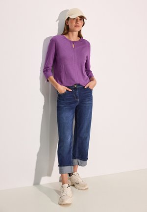 Haut à manches longues violet avec un décolleté en keyhole, associé à un jean large bleu foncé, retroussé au bas, et des baskets beige clair.