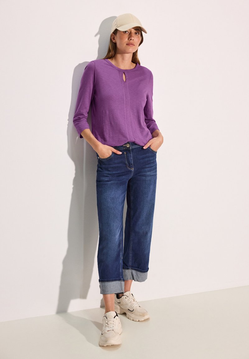 Lila Langarmtop mit einem Ausschnitt in Tropfenform, kombiniert mit dunkelblauen weiten Jeans, die am Saum umgeschlagen sind, und hellbeigen Sneakers.