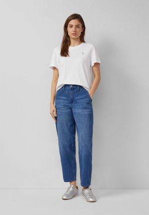 Frau steht vor einer grauen Wand, trägt ein weißes T-Shirt mit einem kleinen Blumendesign, blaue Jeans und silberne Turnschuhe.